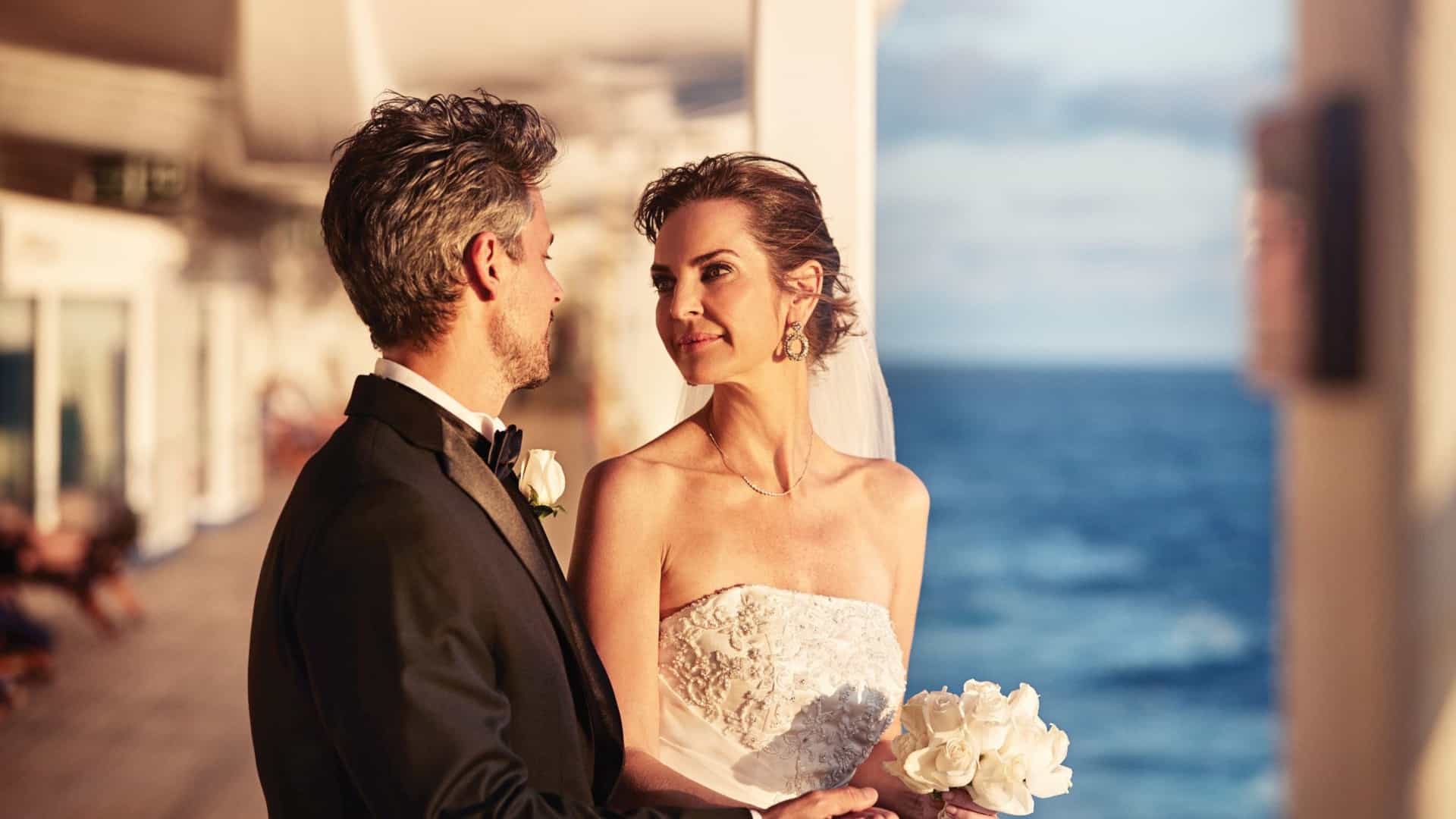 A bride in a strapless wedding dress holding a white rose bouquet shares a romantic look with her groom in a tuxedo on a Princess cruise ship deck at sunset.