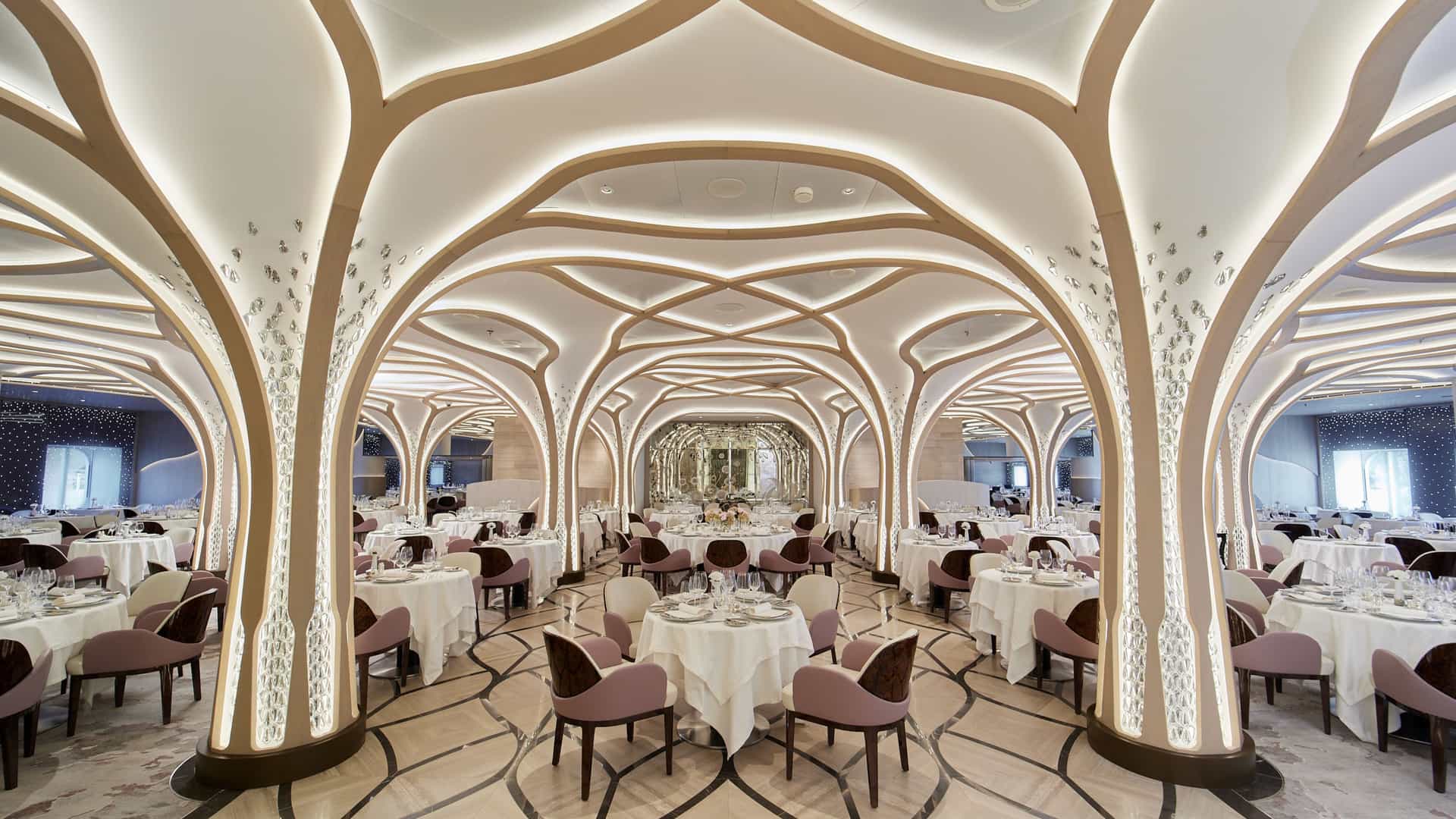 The elegant Compass Rose restaurant aboard the Seven Seas Grandeur features illuminated, tree-like architectural arches, crystal-accented pillars, and rows of tables with mauve chairs.