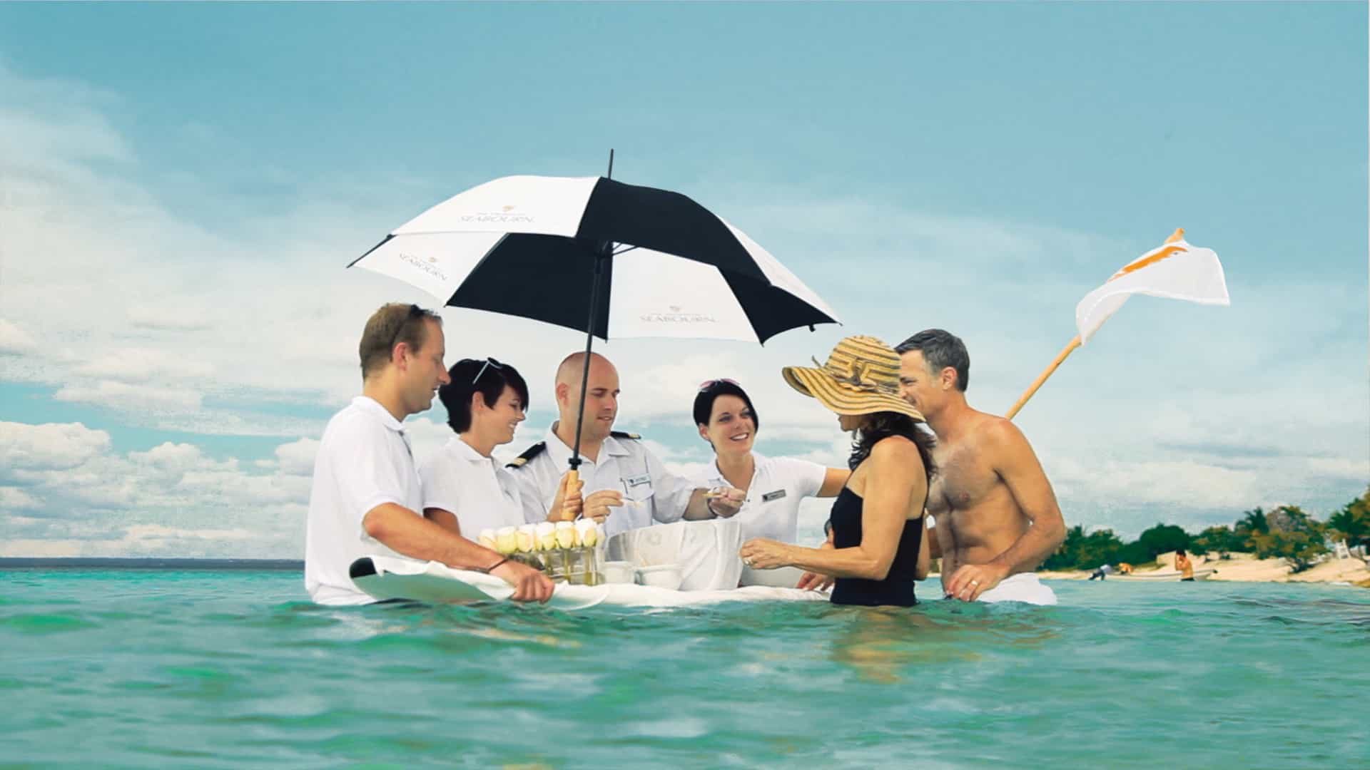 Uniformed Seabourn cruise crew members wading in turquoise ocean water, serving champagne and caviar from a floating white surfboard to a couple during the signature "Caviar in the Surf" beach event.
