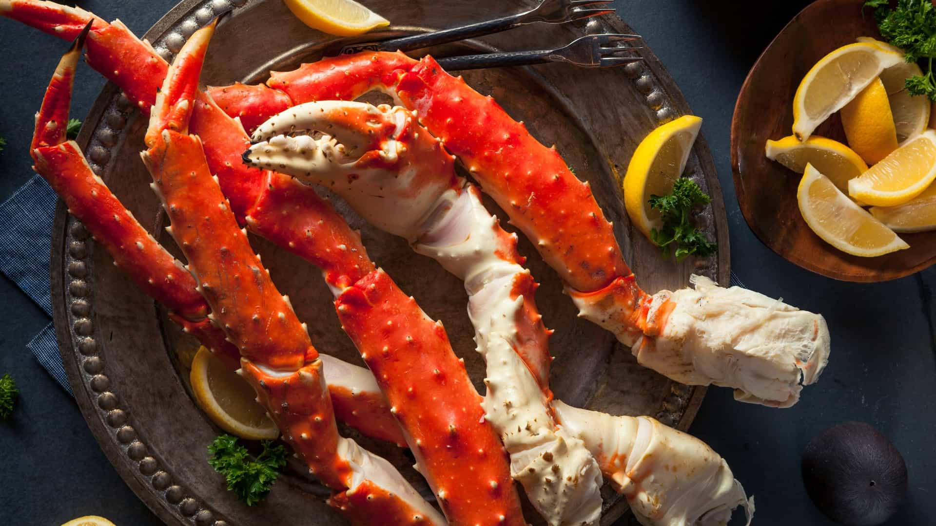 Overhead view of cooked king crab legs and a claw on an ornate round metal tray, with lemon wedges, parsley and two forks, plus a wooden bowl of lemon wedges at the corner.