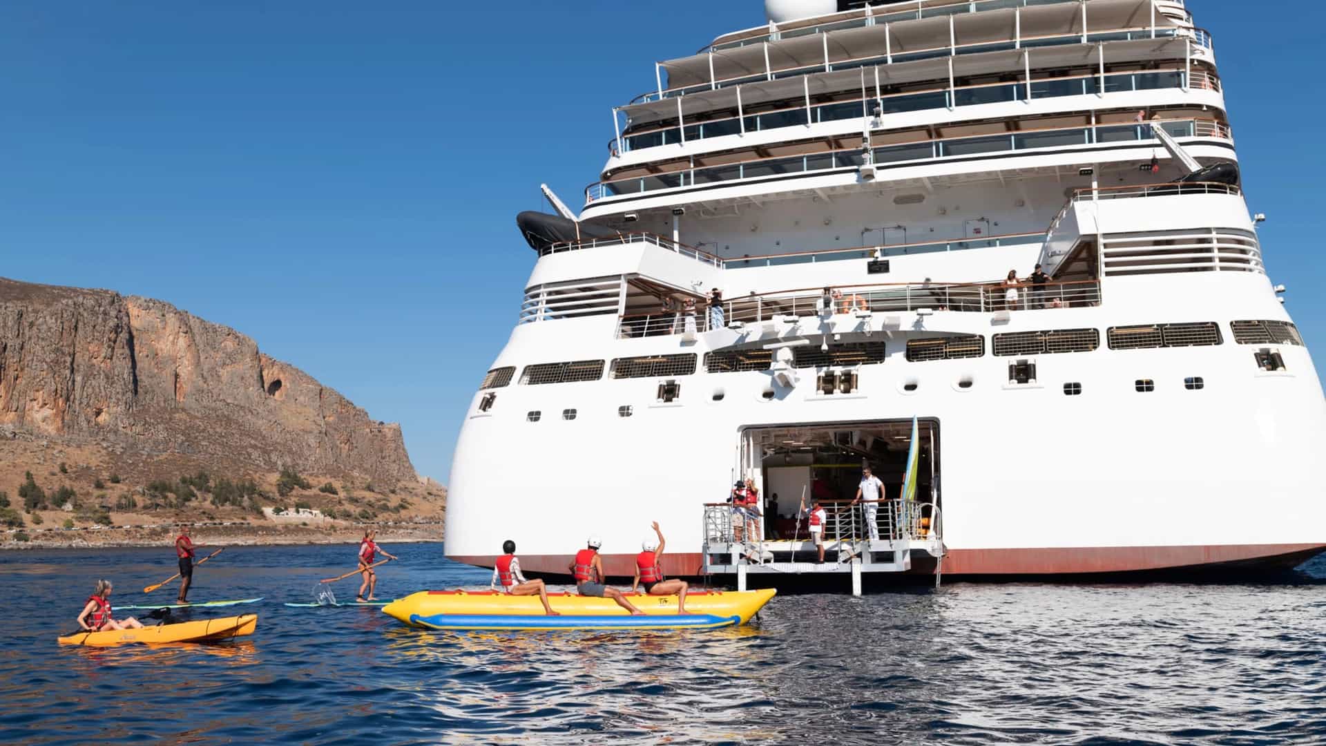 The aft of a white luxury Seabourn cruise ship with its retractable marina platform deployed. Guests in life jackets are participating in water sports, including kayaking, paddleboarding, and a banana boat, against a backdrop of coastal cliffs.