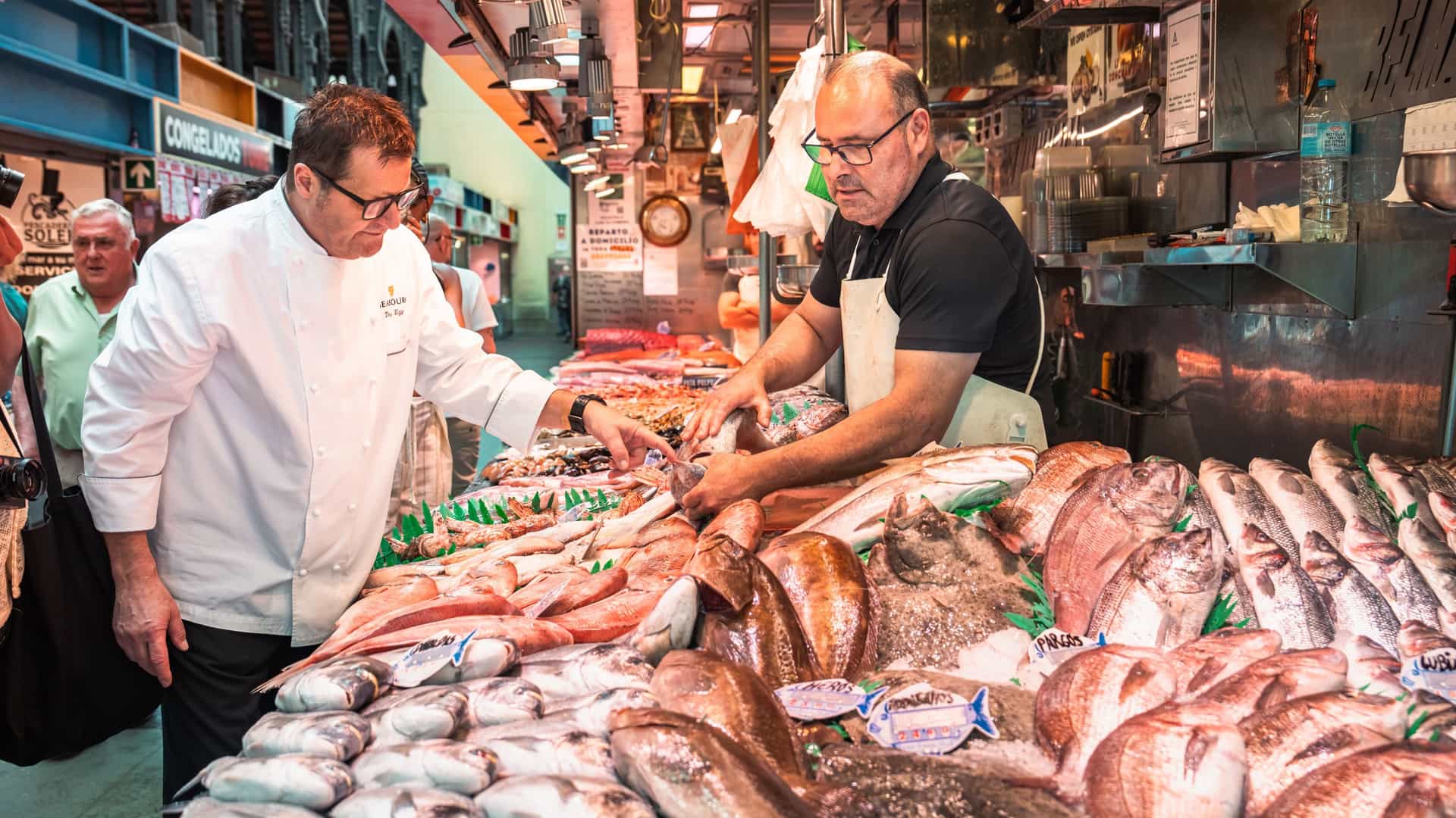 A chef in a white jacket with a Seabourn logo leans over a market fish counter and points at a fish while a fishmonger in a black shirt and apron holds another. Whole fish on ice fill the stall.