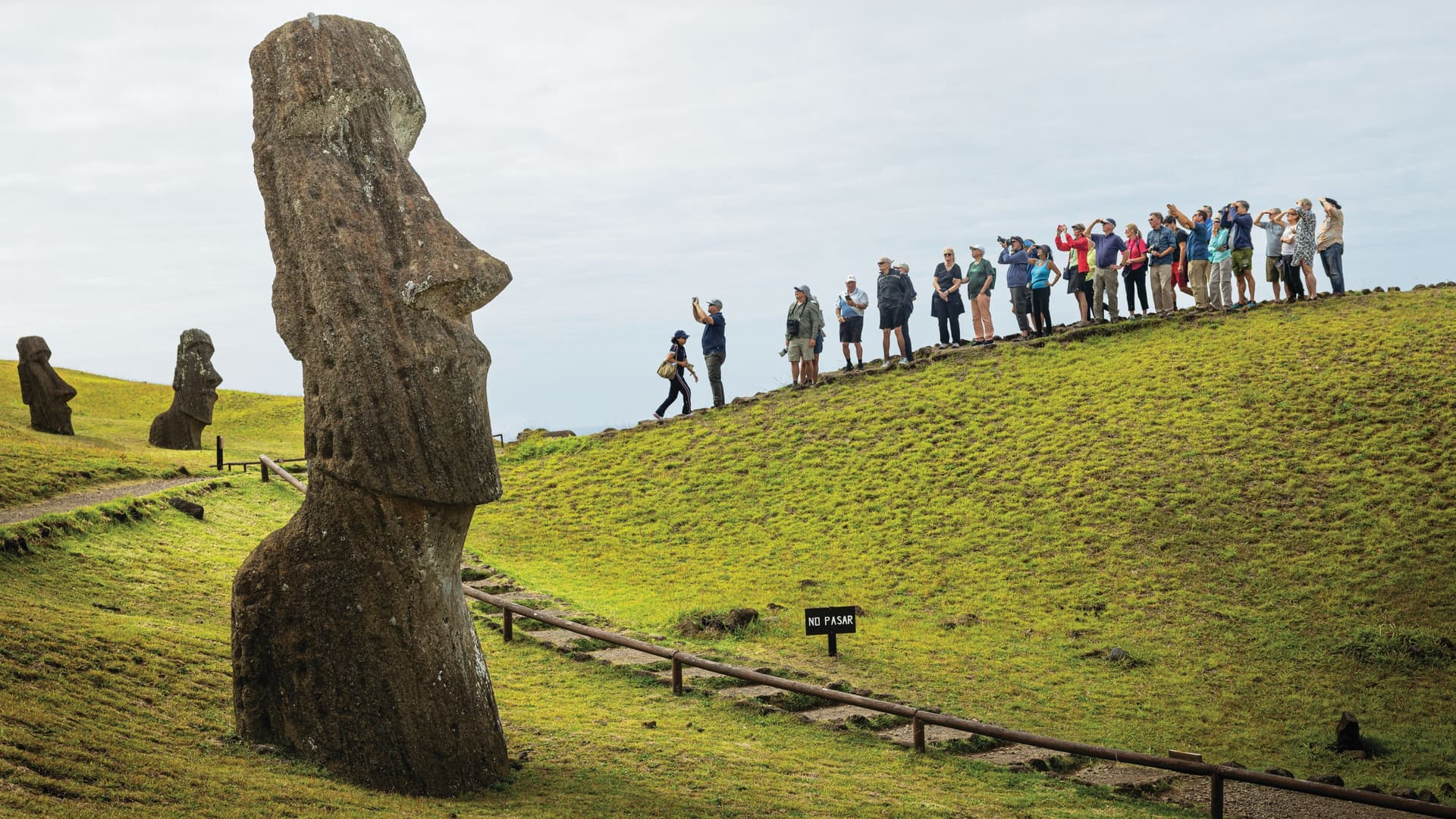 Alt Text Tourists standing along a grassy slope on Easter Island, Chile, viewing monolithic Moai stone statues under an overcast sky.