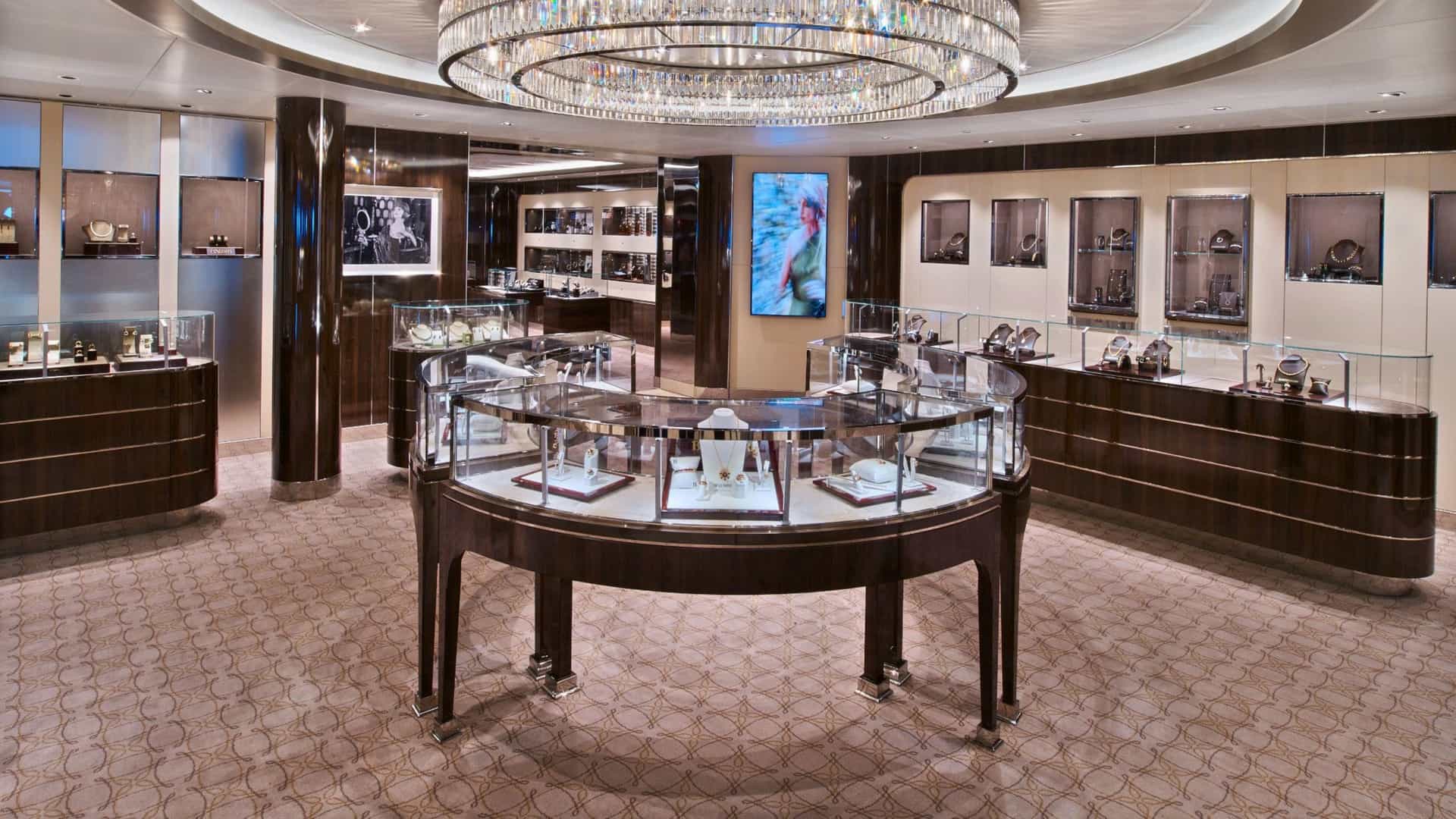 The Boutique on Seabourn Encore featuring glass display cases with luxury jewelry and accessories, dark wood cabinetry, a large crystal chandelier, and patterned carpeting.
