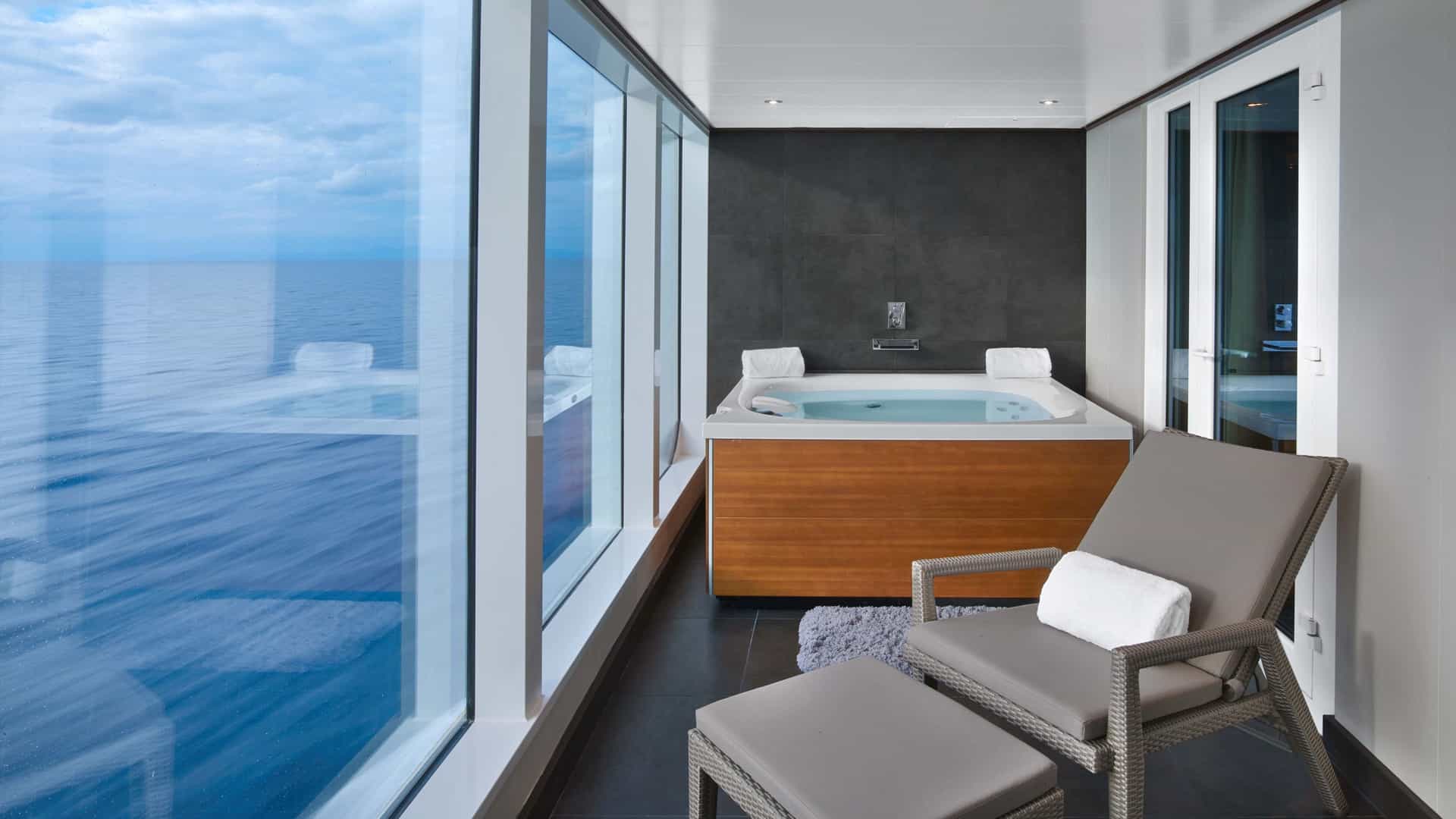 A private glass-enclosed solarium in a Seabourn Encore Wintergarden Suite featuring a wooden jetted bathtub and a lounge chair with panoramic ocean views.