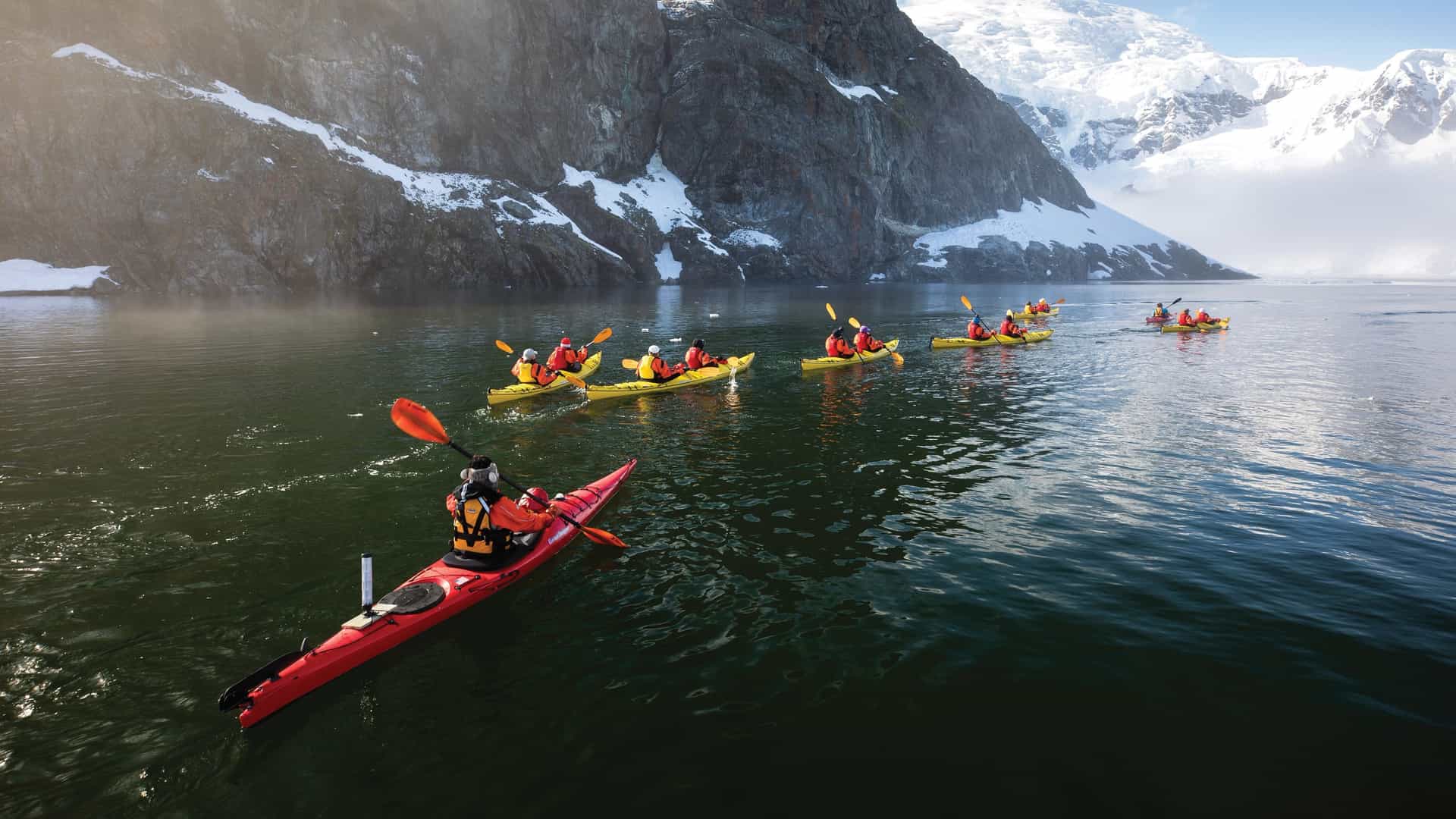 A group of travelers in bright yellow and red sea kayaks paddling through calm, dark water in Antarctica, with massive snow-covered mountains and rocky cliffs in the background..