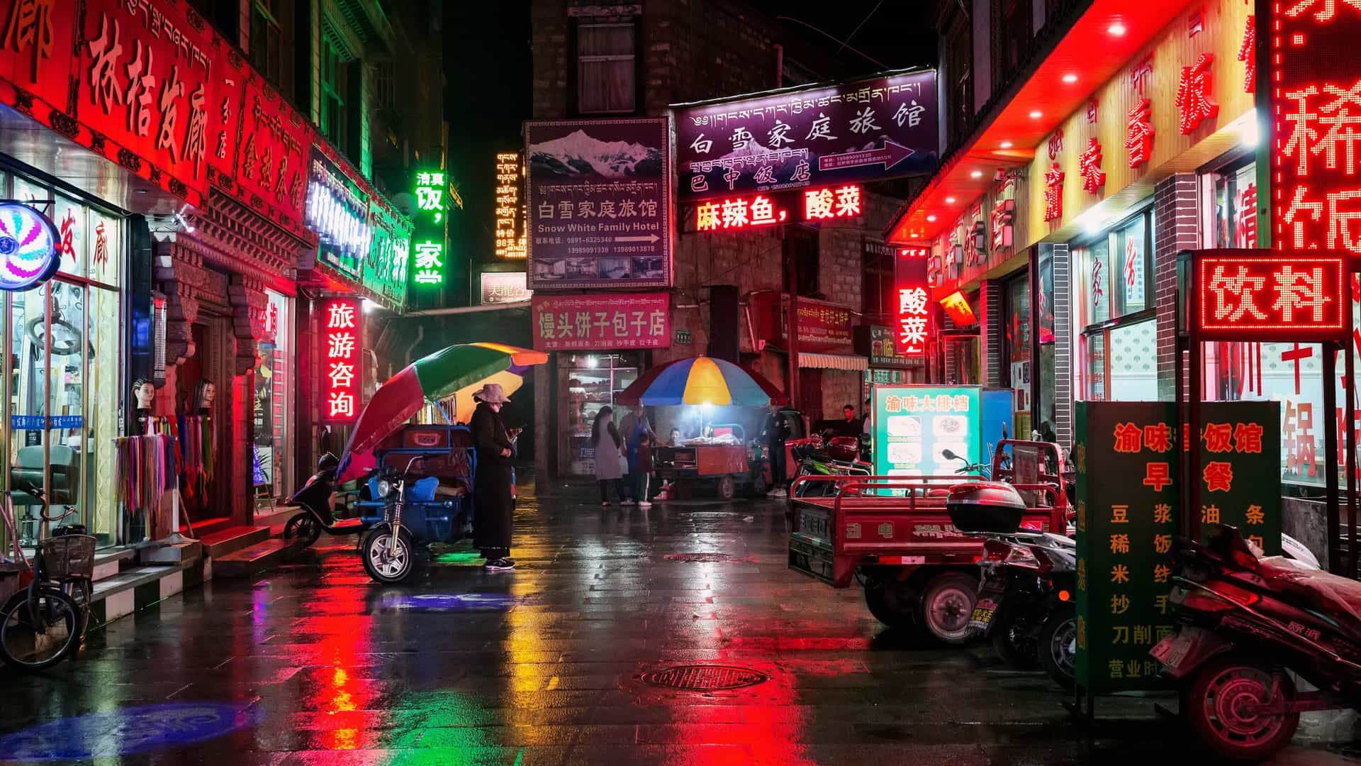 A glowing, neon-lit street scene at night in a Tibetan region, featuring local storefronts, street food vendors under umbrellas, and wet pavements reflecting the bright signs.