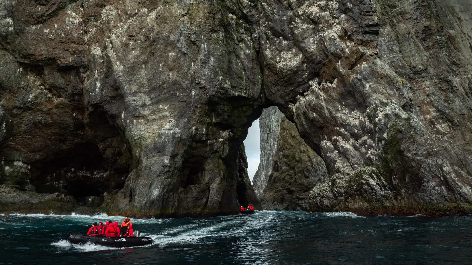 Two Silversea Expeditions Zodiac boats carrying guests in red parkas navigating rough waters beneath towering volcanic sea cliffs and a natural rock arch at Bear Island, Norway.