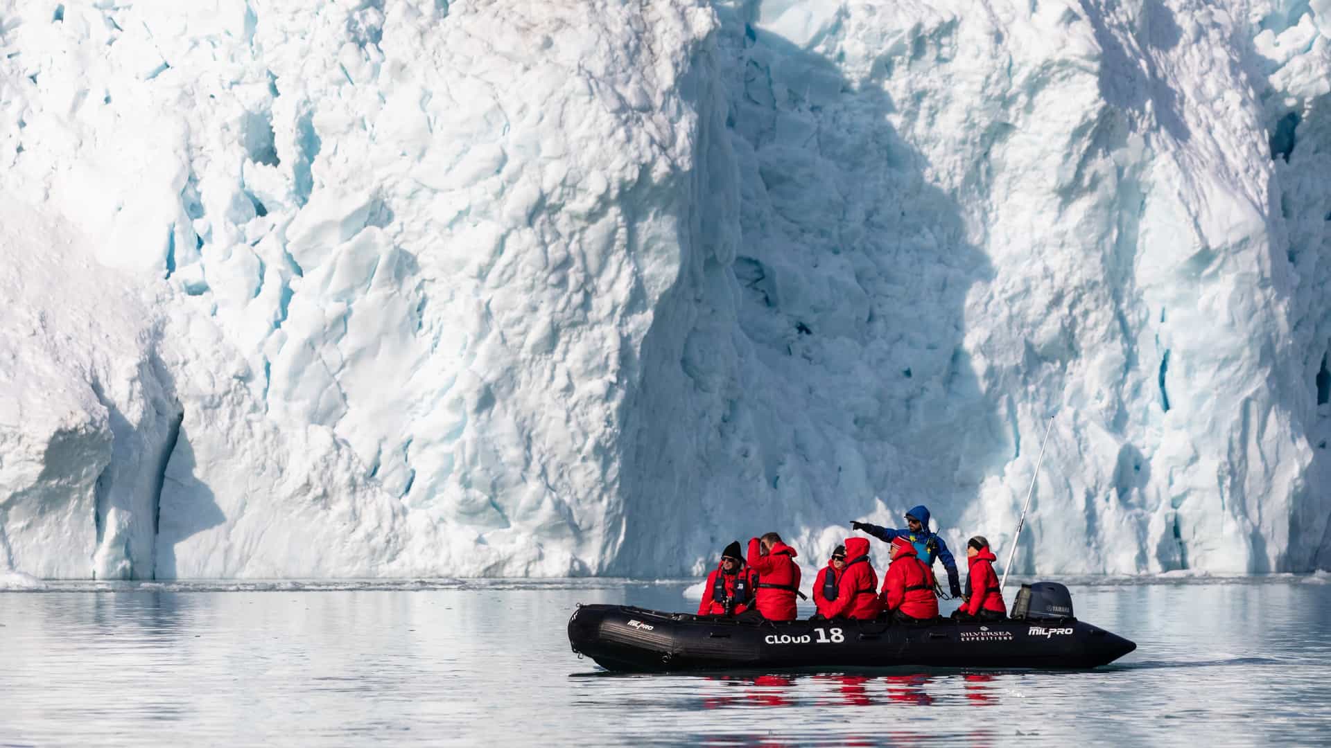 A small group of Silversea Expeditions guests in red parkas aboard a Zodiac inflatable boat, guided by an expedition expert pointing toward a towering glacier wall in Svalbard, Norway.