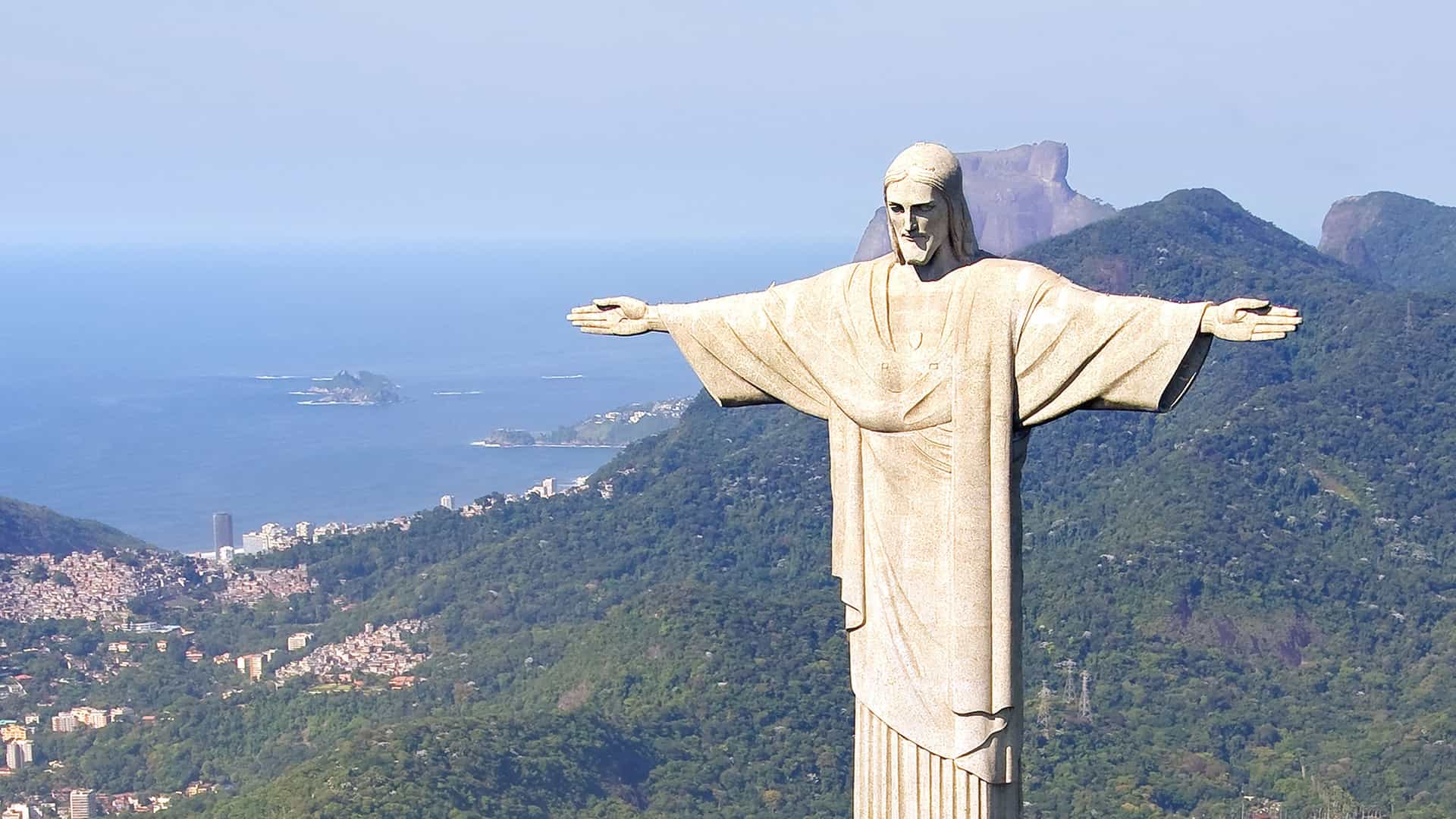 Christ the Redeemer statue stands tall over Rio de Janeiro, Brazil, with arms outstretched against a clear blue sky.