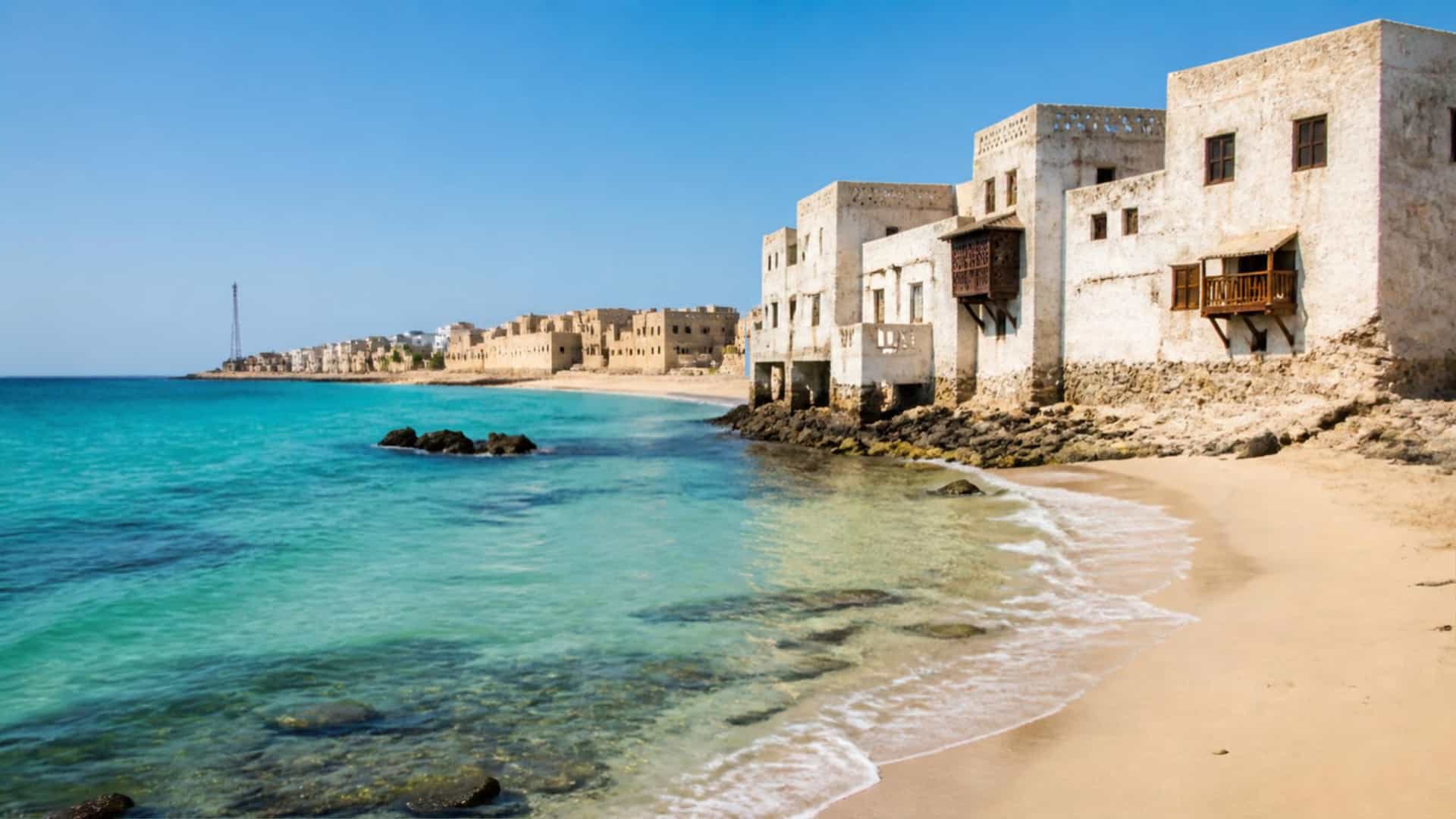 A scenic view of the historic Ottoman-era waterfront in Al Wajh, Saudi Arabia. Whitewashed coral-stone buildings with wooden balconies line a sandy beach, while turquoise Red Sea water laps the shore under a clear blue sky.