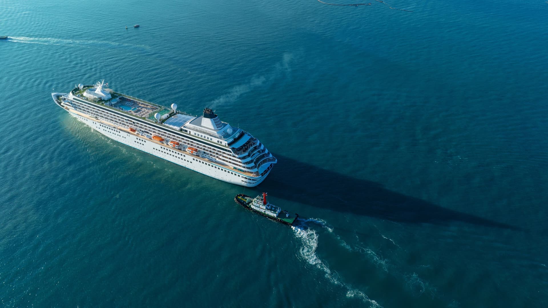 A large white cruise ship sailing across calm open ocean, accompanied by a small tugboat, with sunlight casting a long shadow over the deep blue water as it crosses the equatorial region known as Null Island.