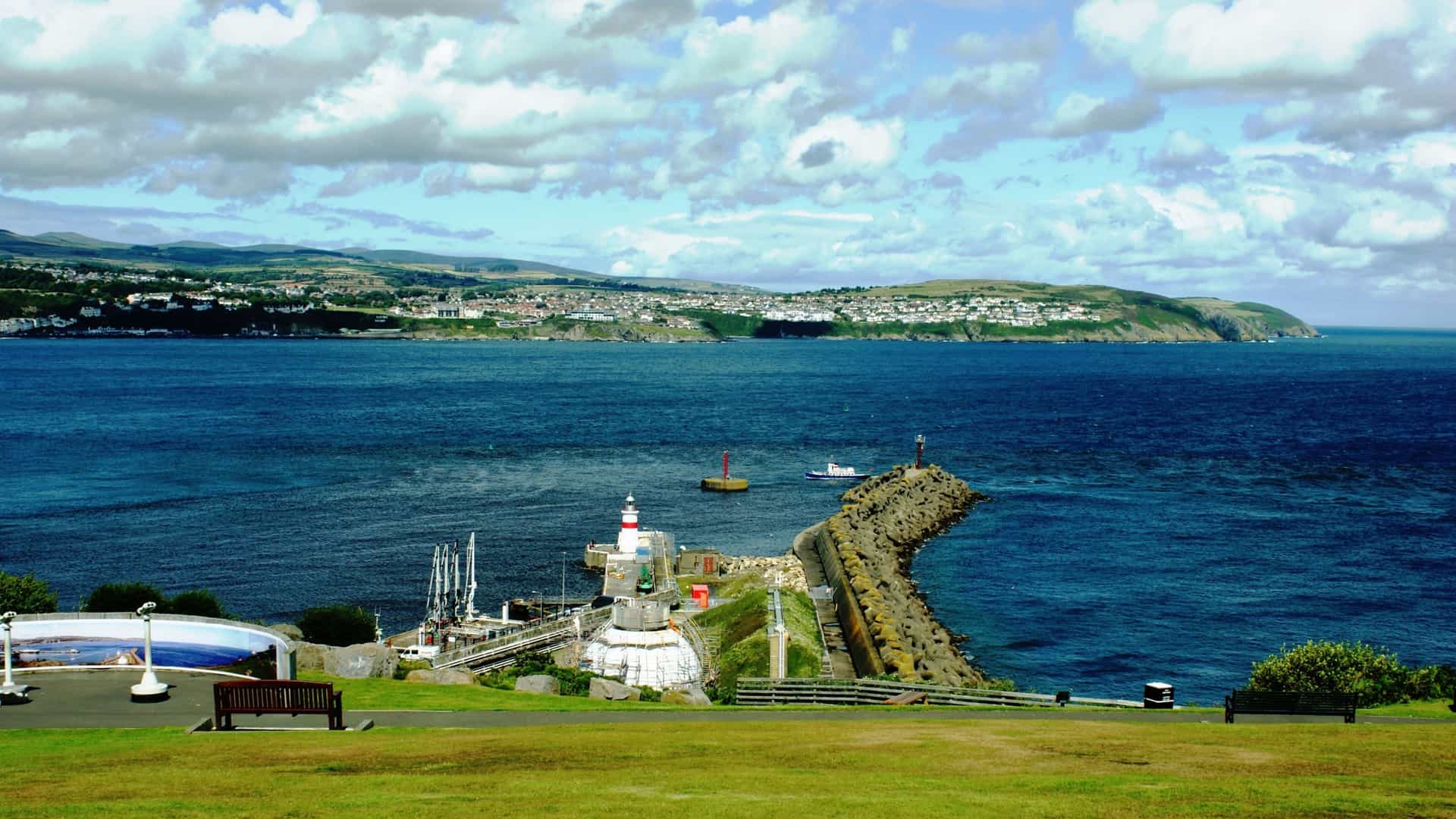 The striped Douglas Harbour lighthouse standing on a curved stone pier, overlooking deep blue water with a distant coastal town and green hills under a partly cloudy sky.