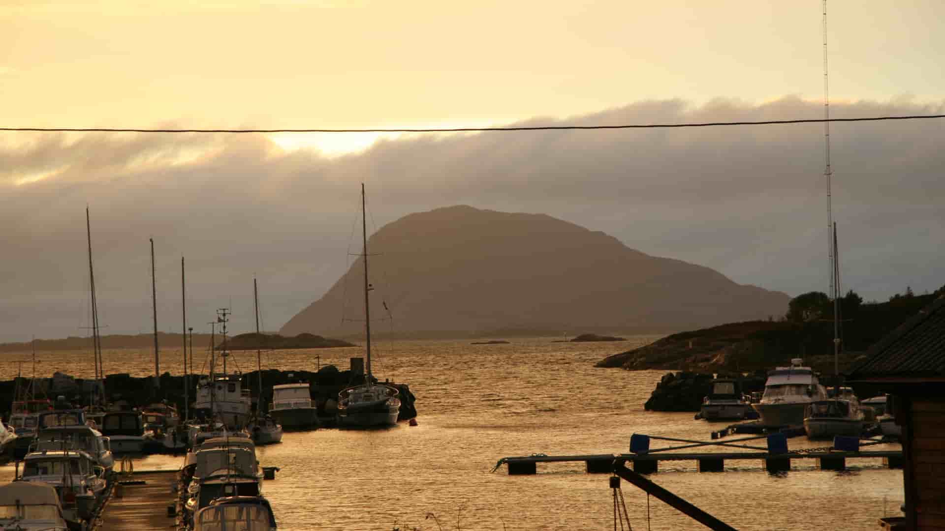 Golden sunset over Florø's harbor in Norway, with fishing boats and sailboats moored in the calm water and forested coastal hills rising behind.
