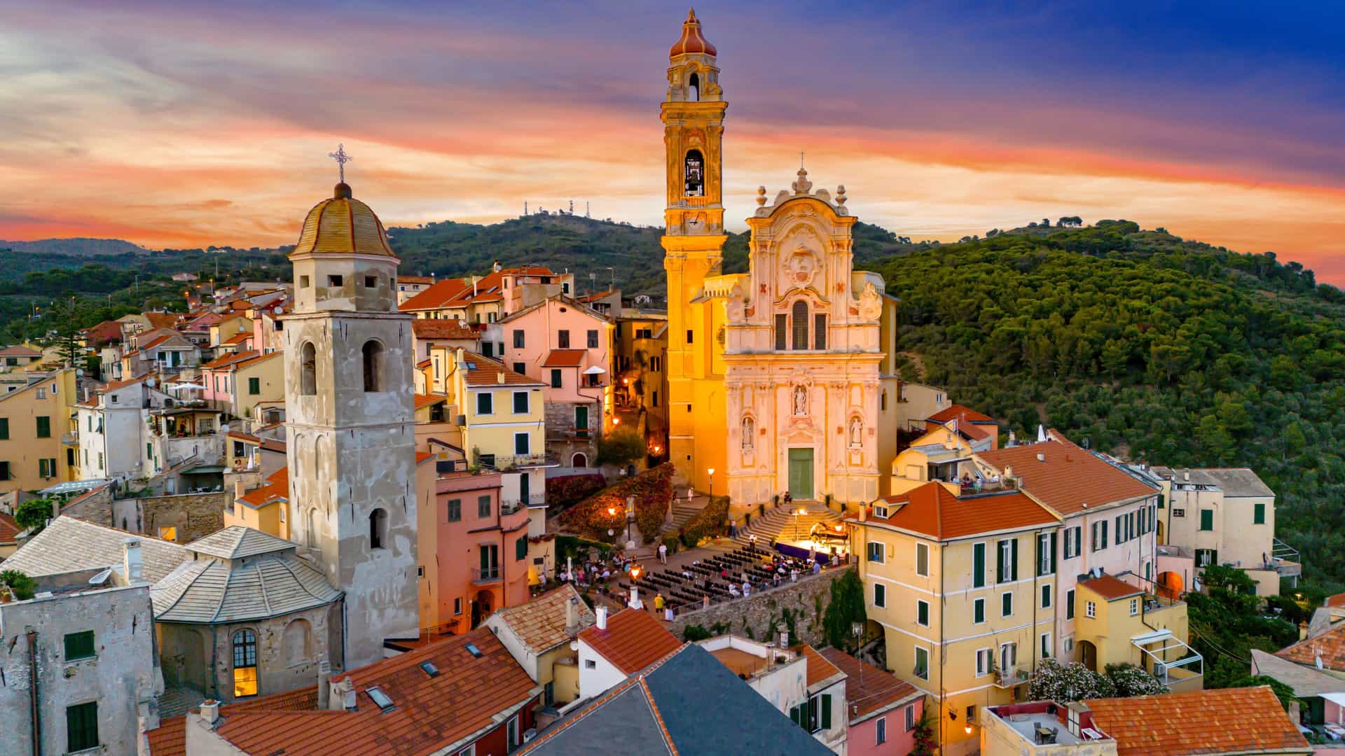 A sunset view of Imperia with colorful hillside buildings, terracotta roofs, and an illuminated Baroque church tower rising above the Ligurian coast under orange‑pink evening skies.