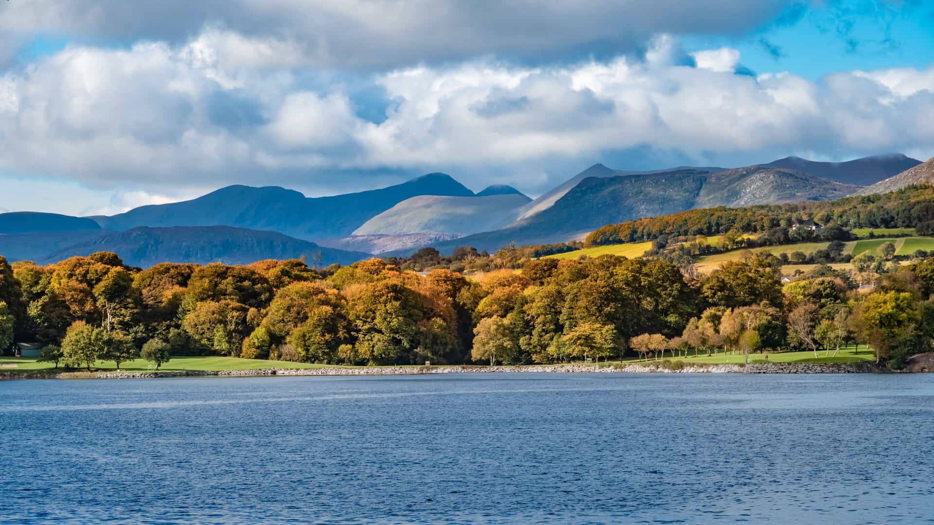 A serene lakeside landscape near Kenmare, Ireland, with calm water, autumn‑colored trees, and rolling mountains under a partly cloudy sky, reflecting the natural beauty of this Ring of Kerry excursion stop.