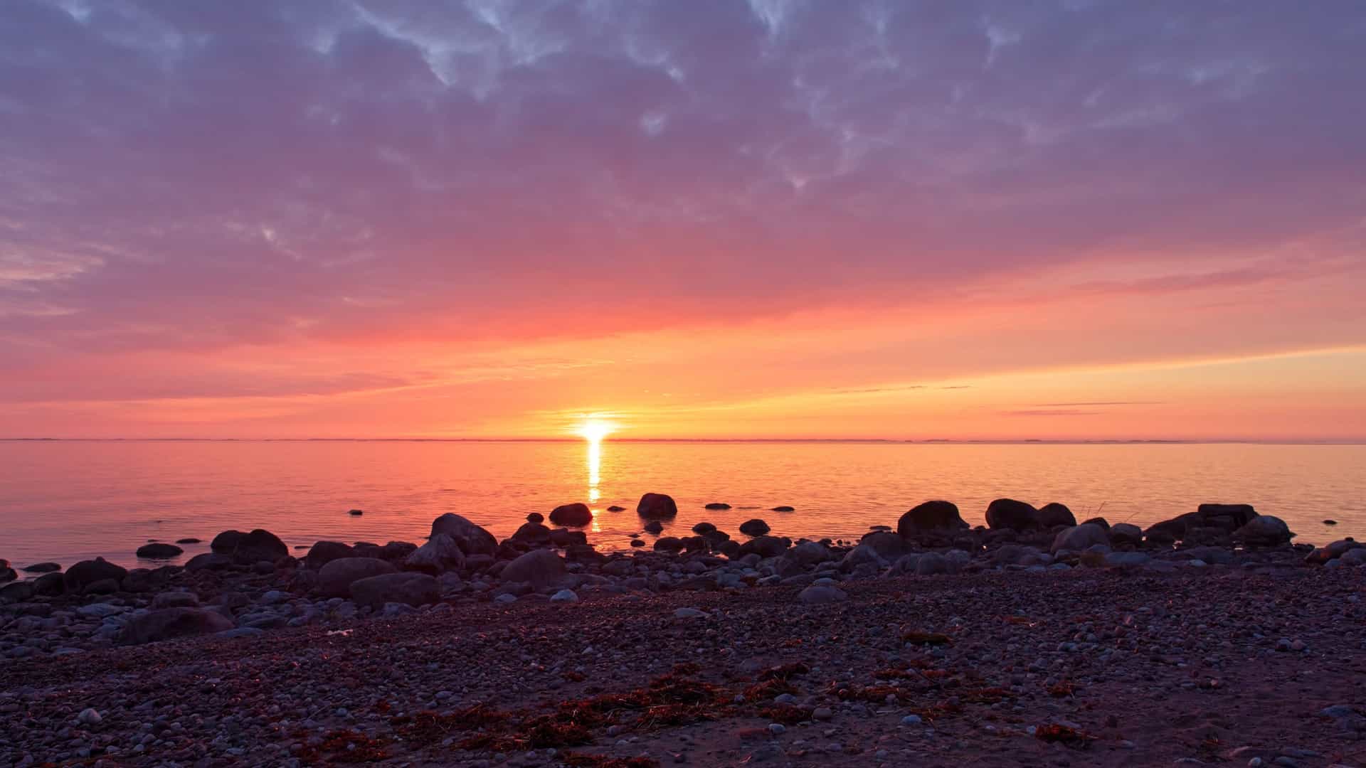 A rocky Oro Island shoreline at sunset, with calm reflective water, scattered stones, and a vivid purple‑to‑orange sky glowing over the Finnish archipelago.