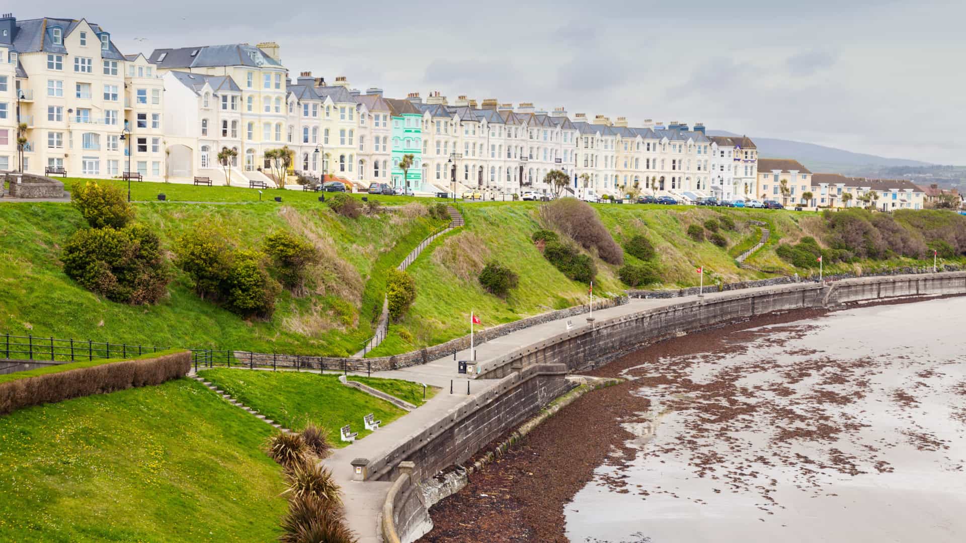 Victorian pastel buildings overlooking the Port St. Mary seafront, with a grassy slope, benches, and a low‑tide beach of wet sand and seaweed under a bright coastal sky.