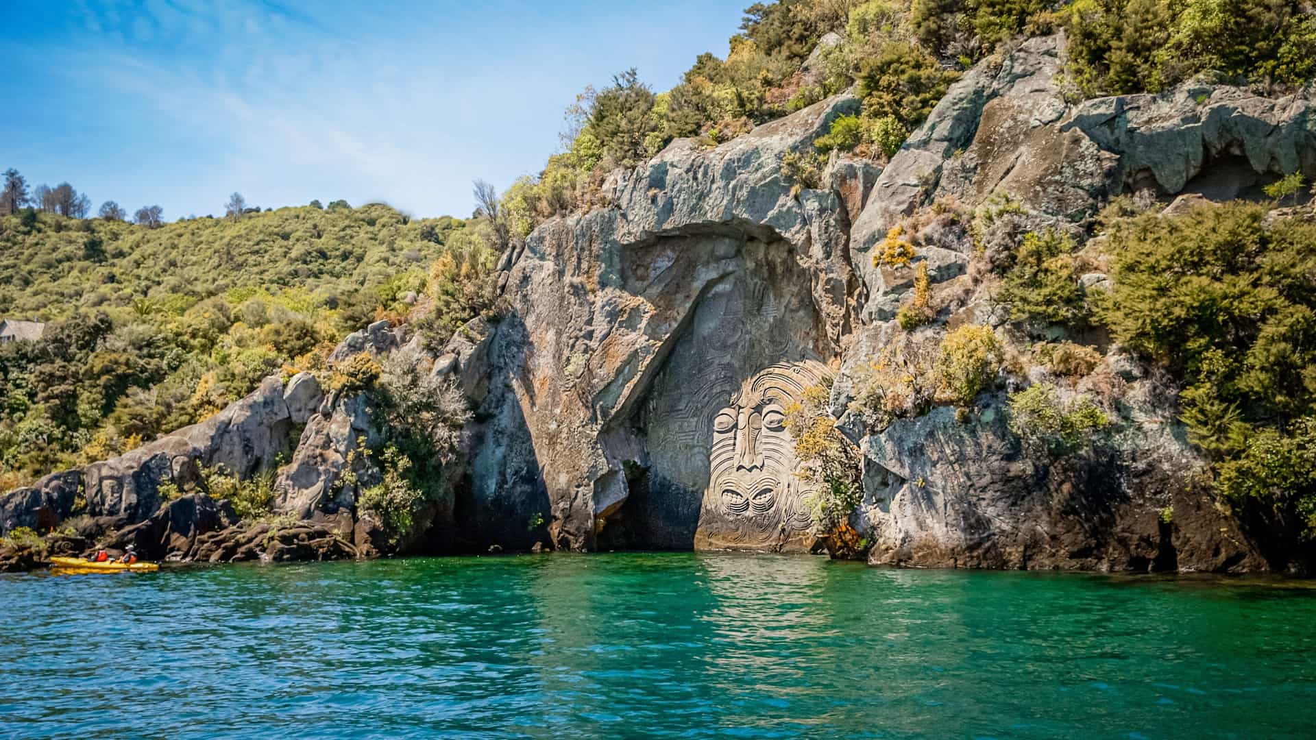The Mine Bay Māori rock carving on Lake Taupō rises from a cliff above turquoise water, depicting a large stylized face surrounded by natural rock and greenery.
