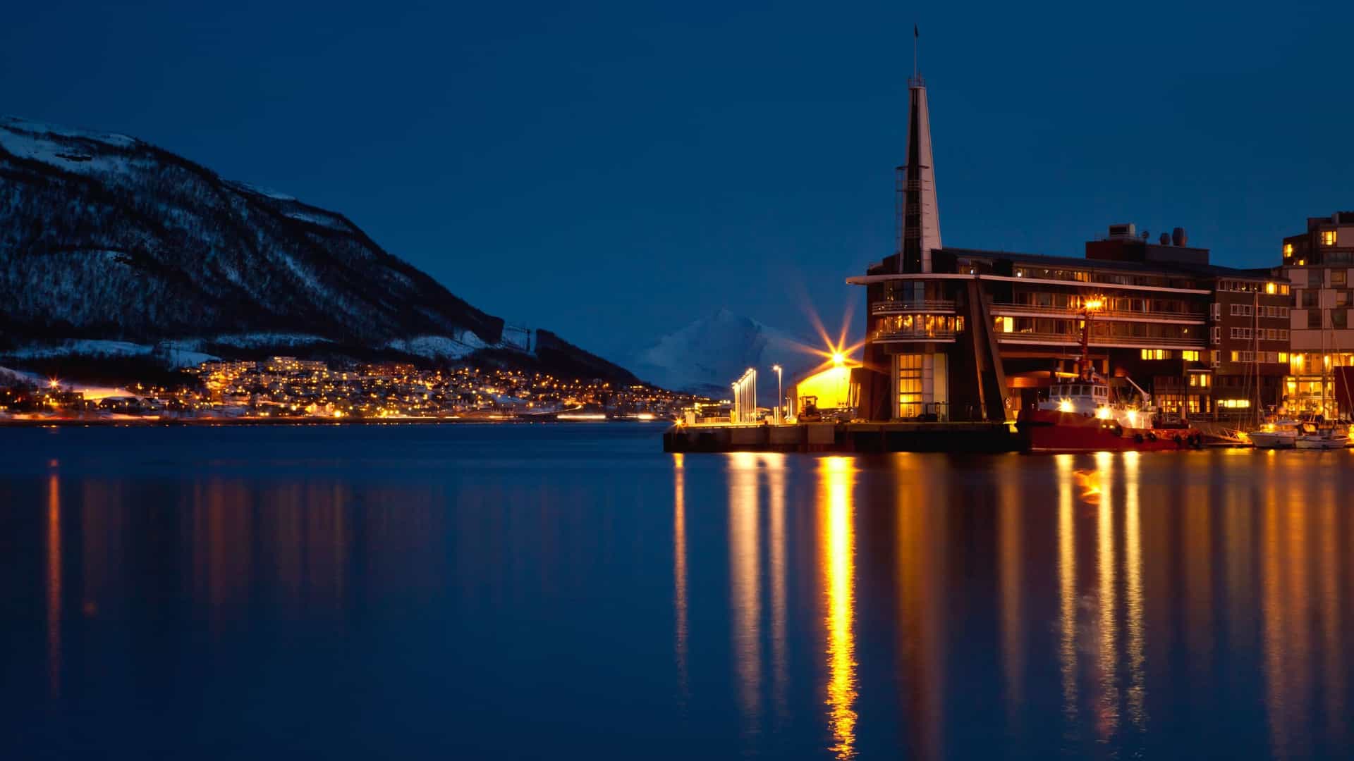 Tromsø harbor at night with illuminated buildings and snowy mountains.