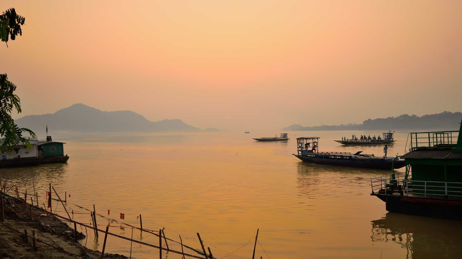 A peaceful scene on the Brahmaputra River in India at sunset, with several small boats on the calm water and mountains in the distance, a destination for a Uniworld river cruise.
