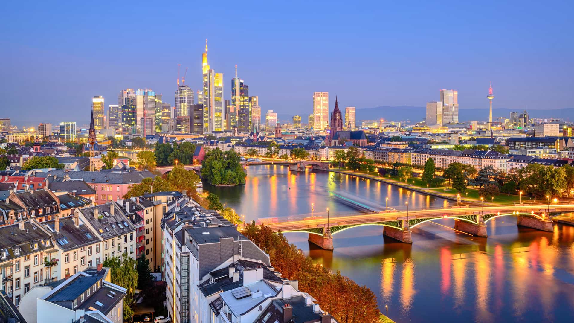 An evening cityscape of Frankfurt, Germany, with the Main River and illuminated bridges flowing past a skyline of modern skyscrapers and historic buildings, a beautiful sight on a Uniworld river cruise.