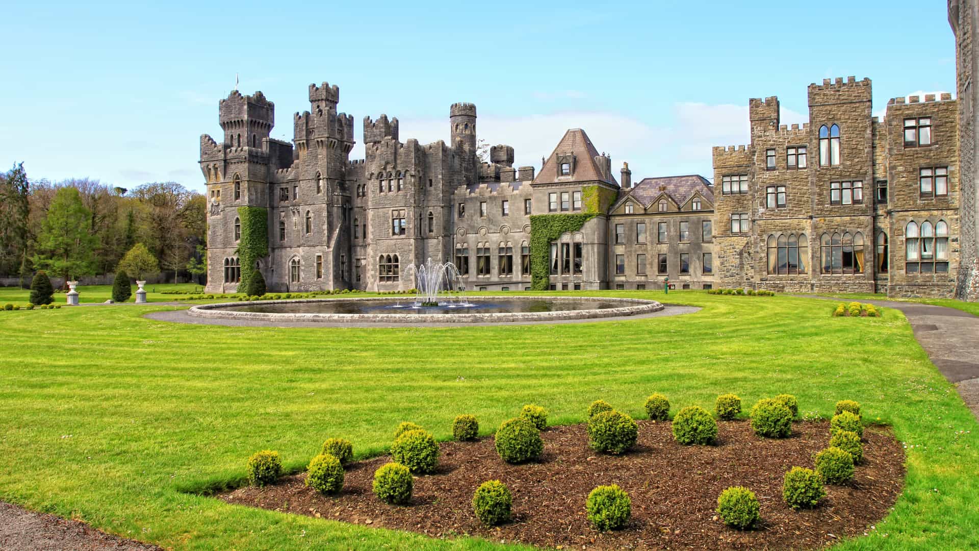 A grand wide shot of the medieval Ashford Castle in County Mayo, Ireland, showcasing its grey stone towers, ivy-covered walls, and a circular fountain centered on a perfectly manicured green lawn.