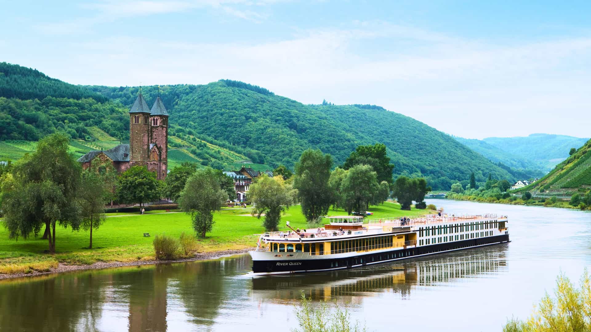 A Uniworld River Queen cruise ship sails on the Moselle River, with rolling hills, vineyards, and a historic stone building on the riverbank under a blue sky, capturing a classic European river cruise scene.