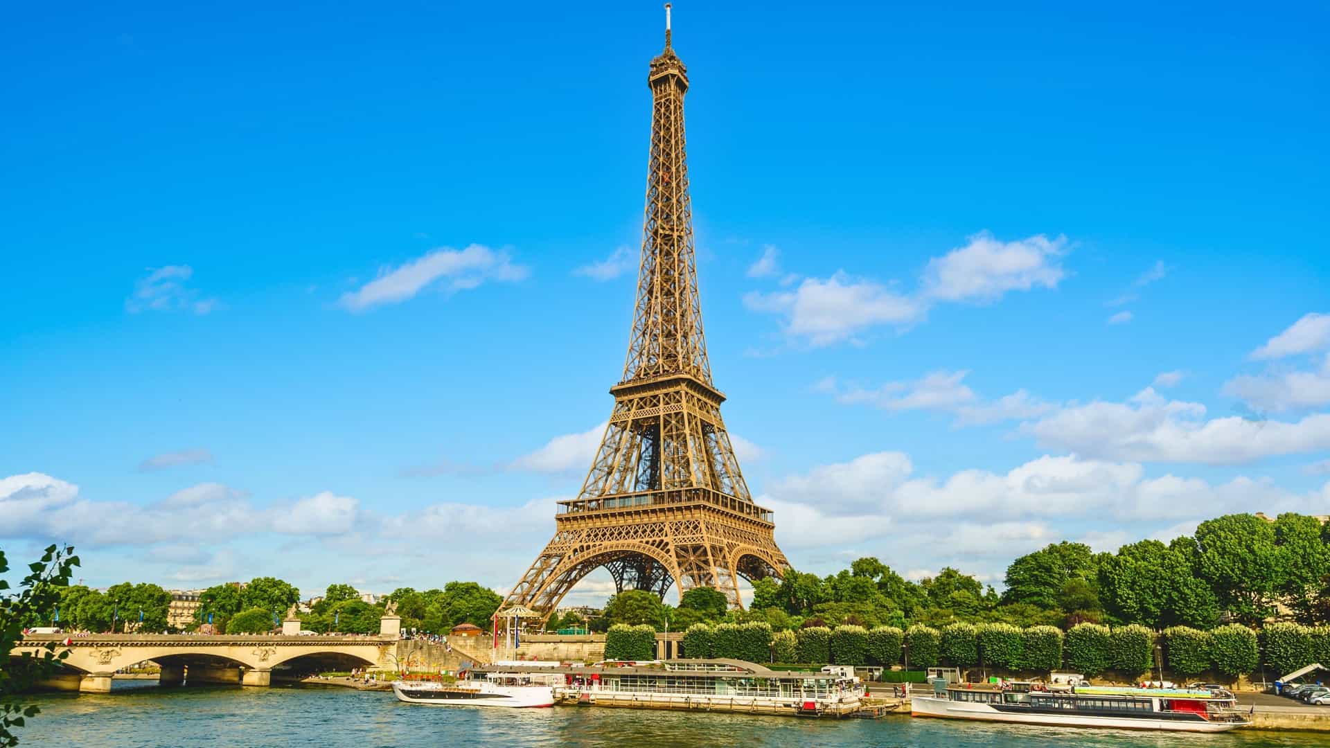 The iconic Eiffel Tower stands tall against a bright blue sky in Paris, France, with the Seine River and Uniworld cruise ships visible in the foreground, a highlight of a French river cruise.