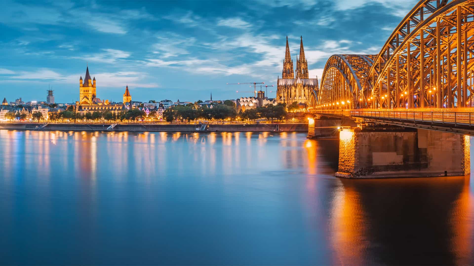 The illuminated Cologne Cathedral and Hohenzollern Bridge at dusk, a stunning view of the Rhine River in Germany and a highlight of a Viking River cruise.