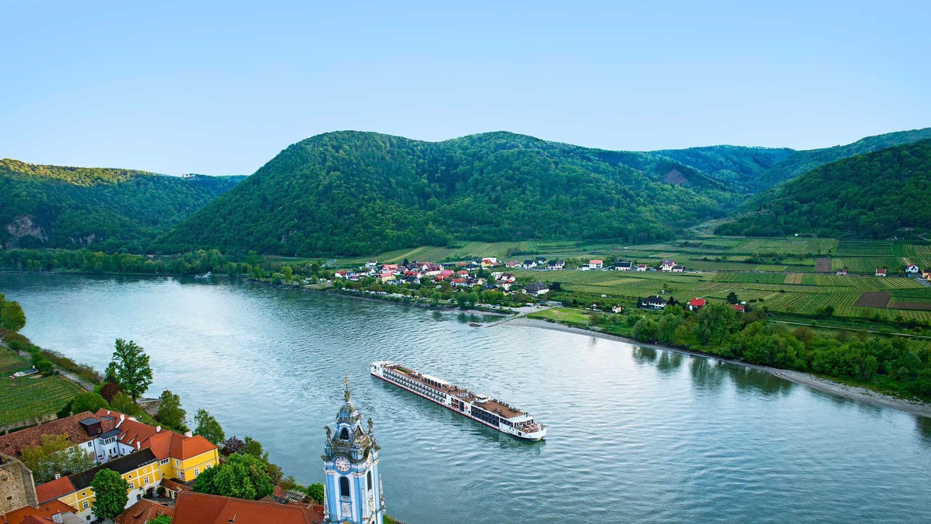 A Viking River cruise ship sailing past the picturesque town of Dürnstein and its iconic, blue-towered church on the Danube River in Austria.