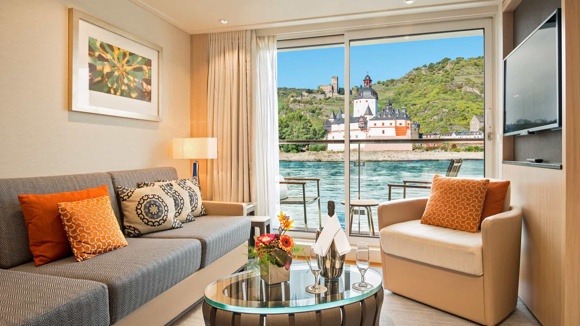 A modern Veranda Suite living area on a Viking river cruise ship features floor-to-ceiling sliding glass doors displaying a scenic view of the historic Pfalzgrafenstein Castle on the Rhine River.