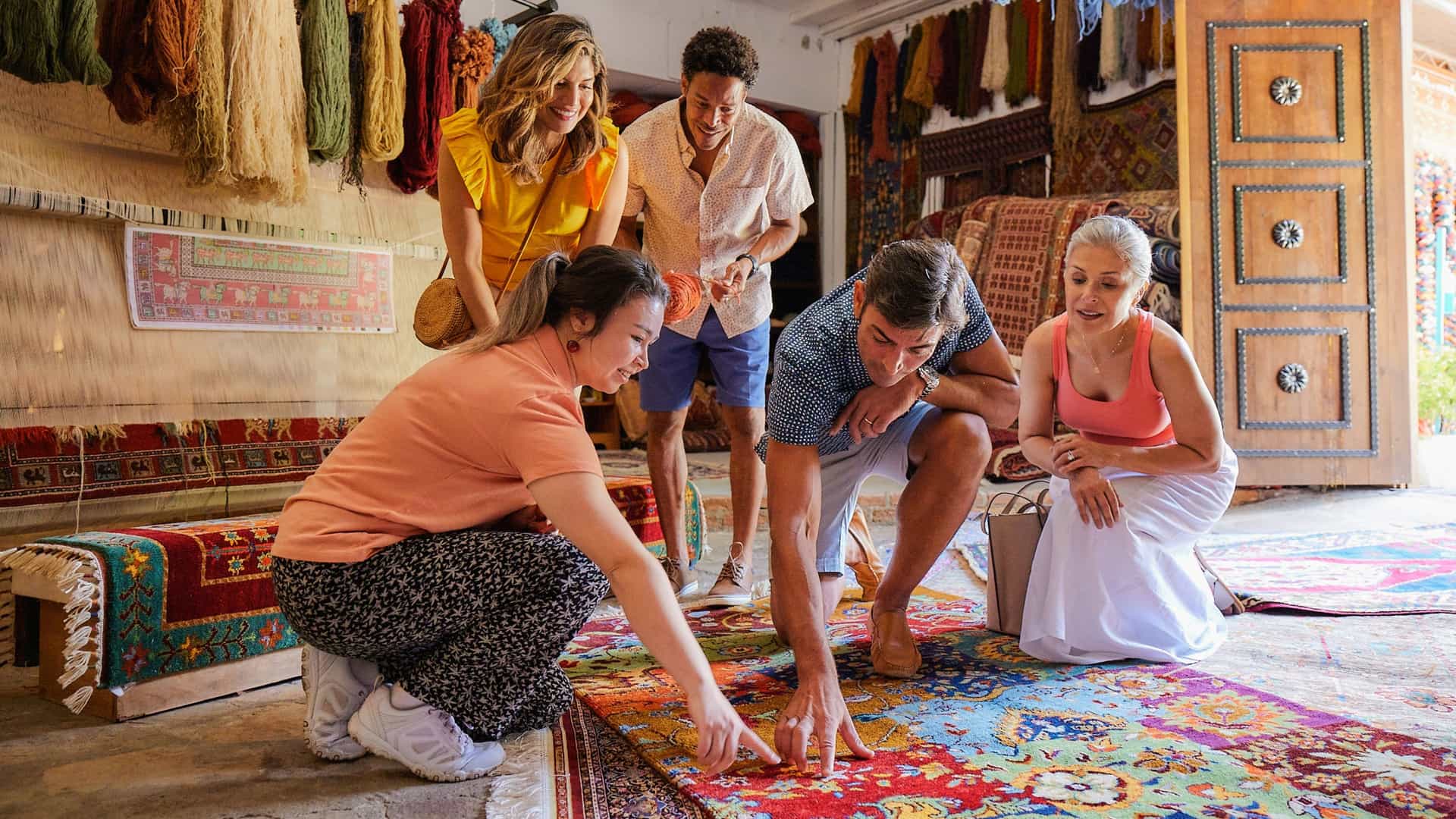 A local artisan shows a vibrant, intricately woven rug to a group of four travelers inside a traditional workshop filled with colorful yarn and textiles.