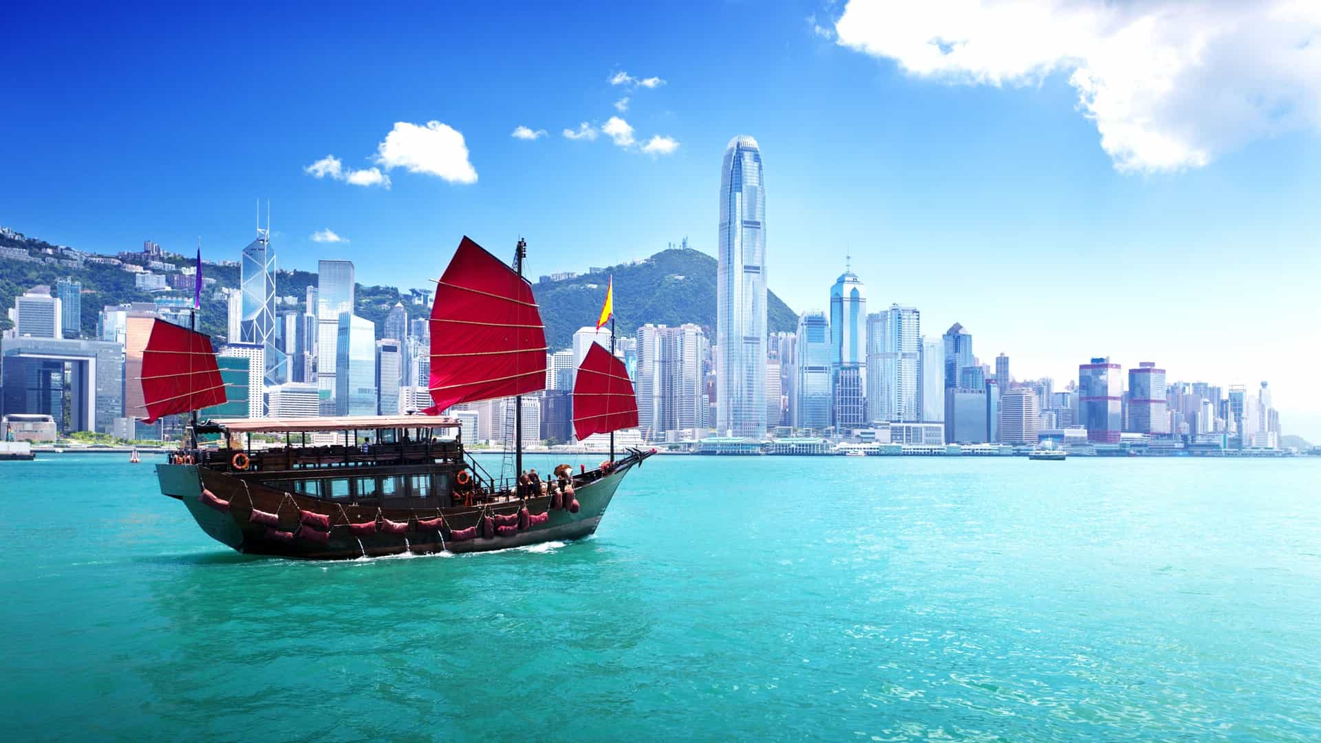 A junk boat with red sails floats on water near a city skyline.